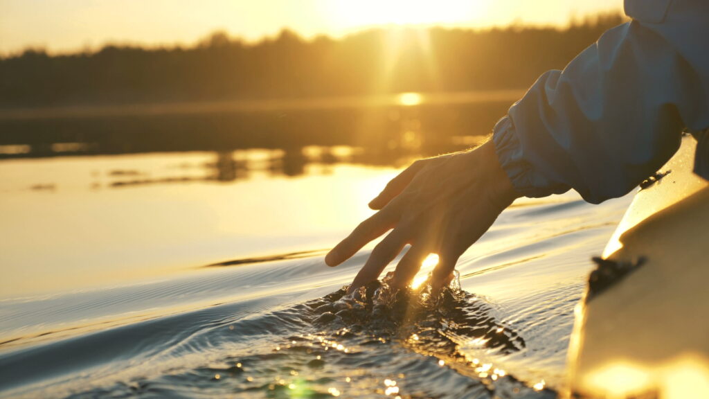 Hand trailing in calm lake water from a kayak at sunset